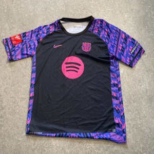 Barça Lamine Yamal Nike fotbollströja L - Snygg FC Barcelona fotbollströja från Nike med Lamine Yamal och nummer 19 på ryggen. Tröjan är svart med lila och rosa grafiskt mönster på ärmarna, rosa detaljer och Spotify-logga på bröstet. Tillverkad i Dri-FIT-material som andas och håller dig sval på planen.