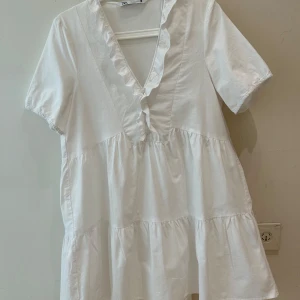  Ny Vit sommar klänning från Zara - Supersöt klänning från Zara med v-ringning och volangkant längs halsen. Klänning har korta ärmar med lätt resår och knappar framtill. Den är luftig med flera volanger i tyget och passar perfekt till ljusa dagar. Gjord i mjuk bomull för en skön känsla.