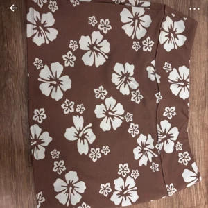 Brun blommig kortkjol - Supersöt brun kortkjol med stora vita blommor i hibiskus-stil. Kjolen har en rak passform och är perfekt för dig som vill sticka ut med ett tropiskt mönster. Skön och lätt att bära, passar perfekt till sneakers eller sandaler.