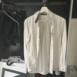 Vit skjorta med mörk krage från Jaguar - Vit långärmad skjorta från nåt Jaguar märke vet inte exakt var  med klassisk passform och mörk kontrastkrage. Skjortan har knappar framtill och en liten broderad logga på bröstet. Tillverkad i bomull, perfekt för en clean och stilren look. Täcken på svarta prickar knappt använd alls storlek 170 motsvarar S