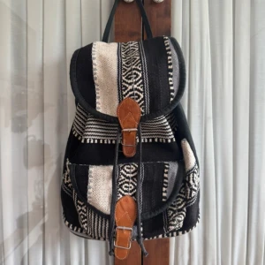 Svart och beige mönstrad ryggsäck - Snygg ryggsäck i svart och beige med etno-inspirerat mönster och detaljer i brunt konstläder. Väskan har två framfickor med spännen och justerbara axelband. Perfekt för dig som vill ha en unik och praktisk väska med coola detaljer.