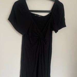 Svart kortklänning med knyt från 157 - Svart kortklänning från 157 med smockad rygg, v-ringning och snygg knytning framtill. Klänningen har korta puffärmar och är gjord i ett mjukt bomullstyg. Perfekt för dig som gillar en enkel men trendig look.
