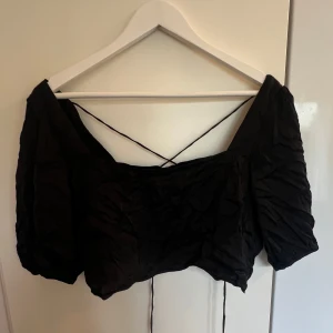 Svart croppad blus/topp med snörning - Svart croppad blus/topp med puffiga korta ärmar och snygg snörning i ryggen. Toppen har en lätt glansig yta och är perfekt till fest, behöver strykas🫣