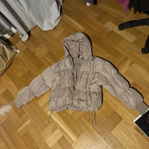 Beige pufferjacka med huva - Säljer en snygg beige pufferjacka med huva. Jackan har dragkedja framtill och justerbara snören i nederkant. Perfekt för kyligare dagar! Den är inte så varm men perfekt för mot sommar tiderna. 