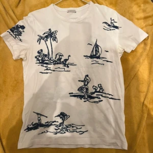 Vit t-shirt med marinblått tryck från Moncler - Säljer en snygg vit t-shirt från Moncler med marinblått tryck av palmer och båtar. T-shirten har korta ärmar och en rund halsringning. Perfekt för en avslappnad stil med en touch av lyx.