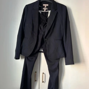 Svart kostym med kavaj och byxor - Elegant svart kostym bestående av en stilren kavaj och matchande byxor. Kavajen har en klassisk design med en knapp och fickor. Byxorna är raka och har en diskret ficka baktill. Perfekt för formella tillfällen.