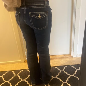 Mörkblå jeans från gina  - Snygga mörkblå jeans med kontrasterande vita sömmar och dekorativa knappar på bakfickorna. Byxorna har en bootcut-stil och låg midja, vilket ger en avslappnad och trendig look. Perfekta för en casual outfit!