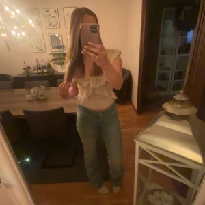 Beige stickad topp med volanger - Säljer en söt beige stickad topp med v-ringning och volangdetaljer. Toppen är ärmlös och har ett fint mönster. Perfekt för att piffa upp vilken outfit som helst!