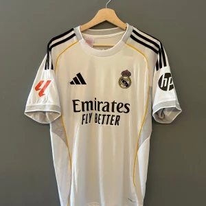 Vit fotbollströja från Adidas 25/26 - Säljer en vit fotbollströja från Adidas med svarta detaljer och gula inslag. Tröjan har korta ärmar och klubbmärke på bröstet. Perfekt för fotbollsfans! Helt ny med tagg i storlek M och en replika