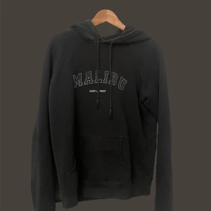 Saint Laurent hoodie  - Säljer en svart hoodie med texten 'Malibu' tryckt på bröstet. Den har en klassisk känguruficka och justerbar huva med snören. Perfekt för en avslappnad stil.