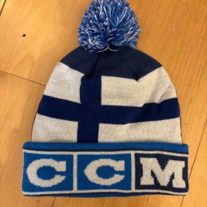 Blå och vit mössa med tofs från CCM - Snygg mössa från CCM i blått och vitt med en stor tofs på toppen. Mössan har ett mönster av en blå korsflagga och texten 'CCM' på uppviket. Perfekt för kalla dagar!