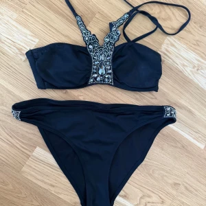 Svart bikini med pärldetaljer från H&M - Snygg svart bikini från H&M med glittrande pärldetaljer på både överdel och underdel. Överdelen har en unik design med pärlor längs axelbanden och en dekorativ mittdel. Perfekt för en stilfull dag på stranden.