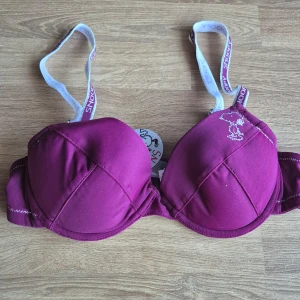 Helt ny bh med Snoopy-design 70A - Snygg vinröd bh med en söt Snoopy-design på kupan. De delvis glittriga axelbanden är justerbara och har Snoopy-text på sig. Uttagbara inlägg. Aldrig använd, prislapp kvar. Kommer från ett konkurslager. Se även mina andra auktioner för möjlighet till bundle med 20% rabatt.