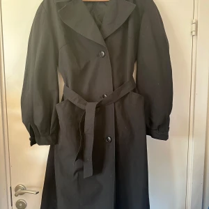 Svart trenchcoat från Distinyo - Snygg svart trenchcoat från Distinyo med knappar och bälte i midjan. Jackan har långa ärmar och en klassisk krage. Perfekt för höst och vår.