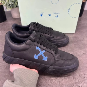 Svarta sneakers från Off-White - Snygga svarta sneakers från Off-White med blått korsbroderi på sidan. Skorna har en robust sula och klassisk snörning. Perfekta för en stilren look.