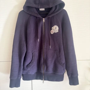 Mörkblå dubble logo hoodie från Moncler - Säljer en snygg mörkblå hoodie från Moncler med dragkedja och dubbel logga på bröstet. Hoodien har en mysig, fleece-liknande textur på framsidan och i luvan,  och justerbar huva med snören. Perfekt för kyliga dagar och kvällar!