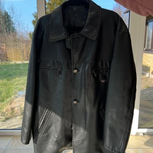 Svart skinnjacka från Avirex - Large - Snygg svart skinnjacka från Avirex med klassisk krage och knäppning framtill, oversized. Jackan har två framfickor med dragkedja och justerbara remmar i midjan för en perfekt passform. Insidan är fodrad med ett mönster av Avirex-loggan.