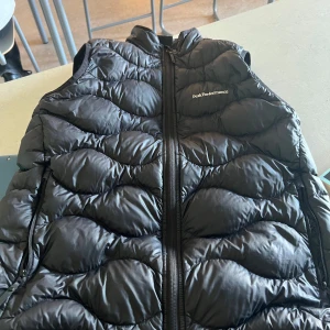 Svart dunväst från Peak Performance - Snygg svart dunväst från Peak Performance med dragkedja framtill. Perfekt för kyliga dagar och ger en sportig look. Västen har en quiltad design som ger extra värme och stil.