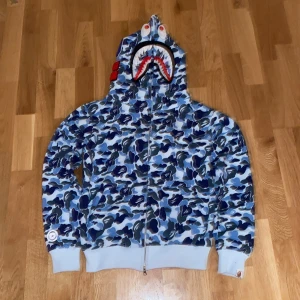 Blå camo hoodie från A Bathing Ape - Säljer en cool blå camo hoodie från A Bathing Ape med hajmotiv på huvan. Hoodien har dragkedja och ribbade muddar. Perfekt för dig som vill sticka ut med en unik stil!