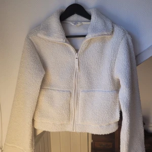 Vit teddyjacka med dragkedja - Mysig vit teddyjacka med dragkedja framtill och två fickor. Perfekt för kyliga dagar med sin fluffiga och varma design. Jackan har en krage och är långärmad, vilket ger extra värme och stil.