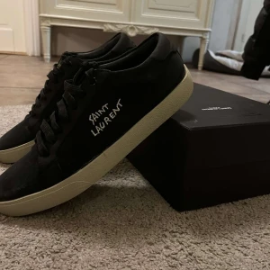 Saint Laurent sneakers svarta - Snygga svarta sneakers från Saint Laurent med vit sula och märkets namn skrivet i vitt på sidan. Skorna har snörning och en klassisk rund tå. Perfekta för en stilren look.