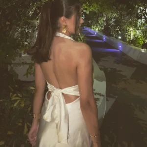 Vit halterneck klänning - Elegant vit halterneck klänning med öppen rygg och knytband i midjan. Klänningen har en v-ringad design och är tillverkad i ett mjukt, flödande material som ger en fin siluett.