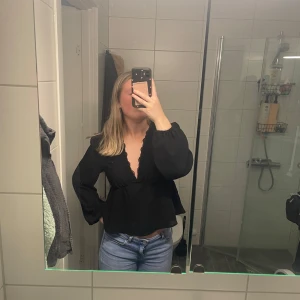 Svart blus med spetsdetaljer - Säljer en elegant svart blus med v-ringning och spetsdetaljer. Blusen har långa ärmar och en lätt rynkad midja som ger en fin passform. Perfekt för en stilren look. Går att pruta. 