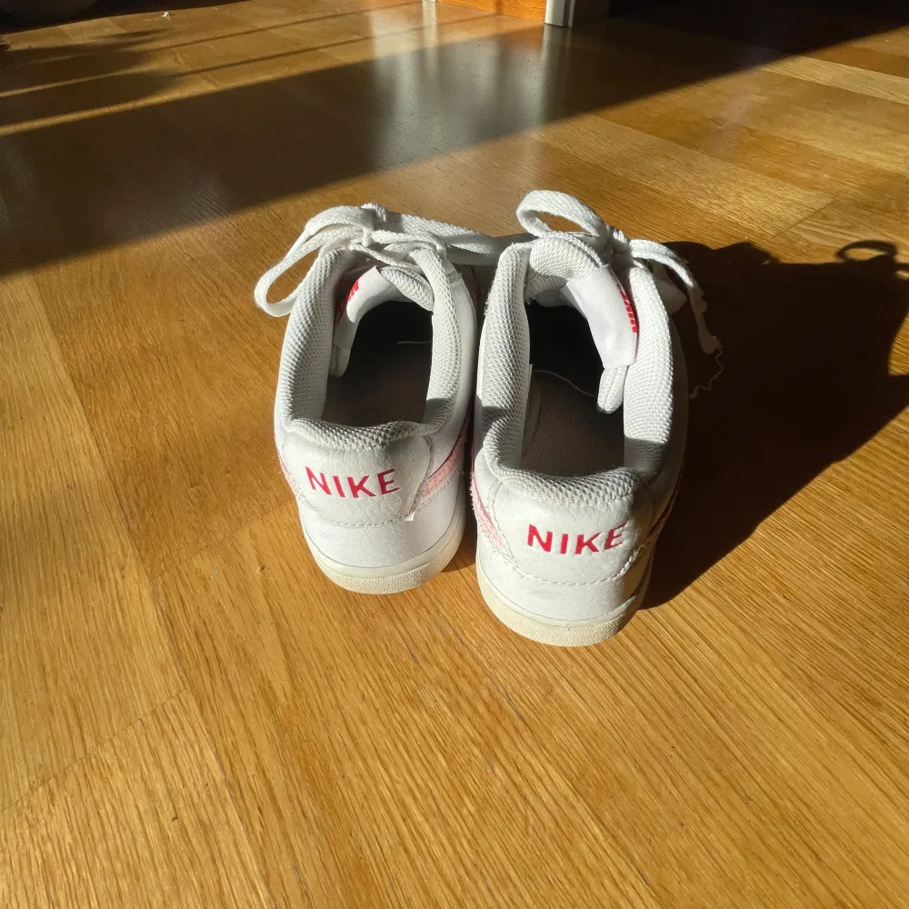 Säljer ett par vita Nike sneakers med rosa detaljer och snörning. Skorna har en klassisk design med perforeringar på tån för ventilation. Perfekta för en avslappnad stil.. Kengät.