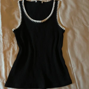 Svart ribbad linnetopp med vit spetskant - Snygg svart ribbad linnetopp med vit broderad kant runt halsringning och ärmhål. Perfekt för en stilren look.