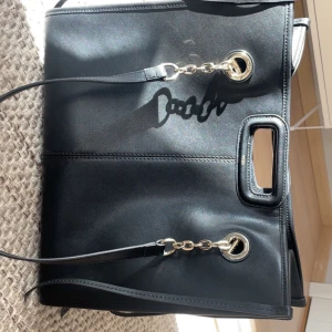 Svart axelväska med kedjedetaljer - Snygg svart axelväska från Maje med kedjedetaljer och praktiska handtag😚. Väskan har en stilren design med metallringar och kedjor som ger en modern touch. Perfekt för att bära dina dagliga nödvändigheter med stil. Pris kan diskuteras vid snabb affär!💕 Ny pris 5500
