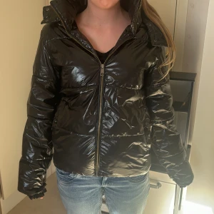 Svart glansig pufferjacka från Nelly - Säljer en svart glansig pufferjacka från Nelly. Jackan har en dragkedja framtill och en hög krage för extra värme. Perfekt för kyliga dagar med sin stilrena look och praktiska design. Lite trasig inne som man ser på sista bilden. Men det syns inte eftersom de är innefrån. Haft den denna vinter och förra vintern, inte så använd. Säljer då jag vill bli av med den och köpa nytt!