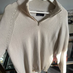 Beige halv zip från ASOS DESIGN - Säljer en stilren beige stickad tröja från ASOS DESIGN. Tröjan har en dragkedja vid halsen och ribbade detaljer vid krage och ärmslut. Perfekt för kyligare dagar! Har en mini fläck på armen som är i princip osynlig. 