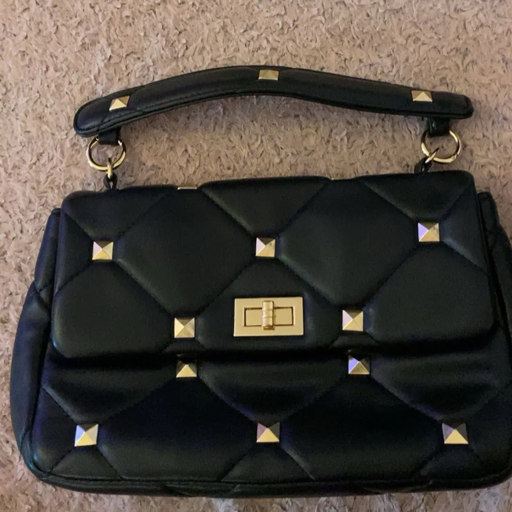 Snygg svart handväska i quiltat mönster med guldfärgade nitar och detaljer. Väskan har ett handtag och en elegant låsdetalj på framsidan. Perfekt för att ge en edgy touch till din outfit. Lite lös vid handtaget. Laukut & Käsilaukut.