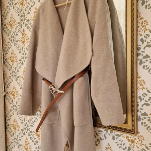 Soft Goat beige kappa - Elegant beige kappa från Soft Goat i en lyxig blandning av 90% ull och 10% kashmir. Den har en stilren design med en bred krage. Storlek L men kan även användas mer oversize av mindre.  Saknar skärpet (läderbälte på bild ingår inte), inga andra anmärkningar 