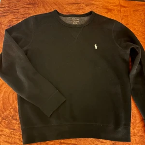 Svart tröja från Polo Ralph Lauren - (NYPRIS 2000kr)Säljer en stilren svart tröja från Polo Ralph Lauren med det klassiska logotypbroderiet på bröstet. Tröjan har långa ärmar och en rund halsringning. Perfekt för en avslappnad och snygg look.