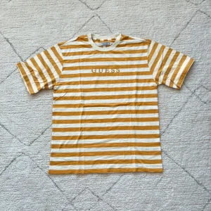 Randig t-shirt från Guess - Säljer en snygg randig t-shirt från Guess i vitt och gult. T-shirten har korta ärmar och ett broderat Guess Los Angeles-tryck på bröstet. Perfekt för en avslappnad stil!