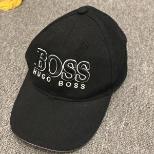 Svart keps från Hugo Boss - Snygg svart keps från Hugo Boss med broderad logotyp framtill. Justerbar rem baktill med metallspänne för perfekt passform. Perfekt för en stilren look.