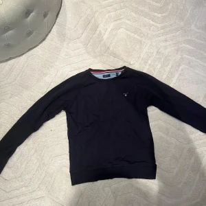 Mörkblå tröja från GANT - Säljer en stilren svart tröja från GANT med rund halsringning och diskret logga på bröstet. Tröjan har långa ärmar och ribbade muddar vid ärmslut och nederkant. Perfekt för en avslappnad look. 