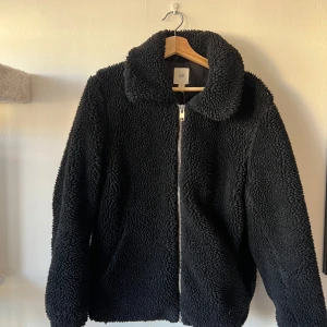 Svart teddyjacka från H&M - Mysig svart teddyjacka från H&M med dragkedja framtill. Perfekt för kyliga dagar med sin fluffiga och varma design. Jackan har en klassisk krage och långa ärmar.