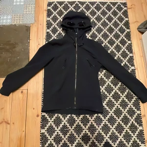 Mörkblå jacka med huva och glasögon från C.P. Company - Säljer en mörkblå jacka från C.P. Company med unik design. Jackan har en huva med inbyggda glasögon och dragkedja framtill. Perfekt för en stilren och modern look. Pris kan diskuteras ny pris 4500kr