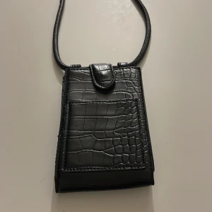 Svart mobilväska med krokodilmönster - Mobilhållare/mobilväska. Plats för en mobiltelefon o kort o läppglans. Höjd 14 cm, bredd: 9 cm o ca 3 cm djup. 1 litet ytterfack. Inga fler fack inuti utan bara för mobilen o något mer småt typ kort/nyckel/läppglans