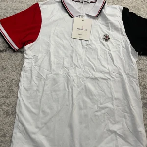 Vit pikétröja från Moncler - Säljer en stilren vit pikétröja från Moncler med kontrasterande röda och svarta ärmar. Tröjan har en klassisk krage med röd och svart randig detalj samt Monclers logotyp på bröstet. Storlek M Men passar L 