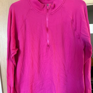 Rosa långärmad tröja med half zip strl: XL - En starkt rosa långärmad tröja med half zip framtill och tumhål i ärmsluten. Tröjan är ribbad och har en sportig känsla, perfekt för lager-på-lager eller träning. Materialet är mjukt och stretchigt, vilket gör den bekväm att bära under kyligare dagar. Förekommer endel noppor vid sidan på tröjan.
