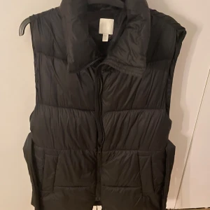 Svart dunväst med dragkedja - Svart dunväst med hög krage och praktiska fickor framtill. Västen har en quiltad design och stängs med dragkedja. Perfekt att slänga över en hoodie för extra värme under kyliga dagar.Den är lång 