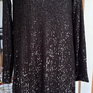 Svart glittrig kortklänning från Amisu - Svart kortklänning från Amisu med långa ärmar och djup v-ringning. Klänningen är täckt av glittriga paljetter som ger en riktigt festlig vibe. Materialet är mjukt och följsamt, perfekt för att sticka ut. Passformen är normal och klänningen har en snygg siluett.