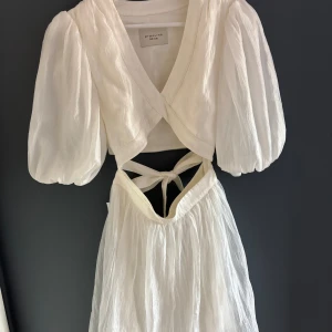 Vit omlottklänning By Malina Bridal - Supersöt vit klänning  från By Malina Bridal med puffiga korta ärmar och v-ringning. Klänningen har öppen rygg med knyt och är tillverkad i ett skirt, glansigt material som ger en lyxig känsla. Perfekt för dig som vill ha en romantisk och trendig look.