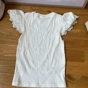 Vit topp med spetsärmar från Gina Tricot - Supersöt vit topp från Gina Tricot med ribbad struktur och korta, volangiga spetsärmar med broderade detaljer. Perfekt för dig som gillar en romantisk och trendig stil. Rund halsringning och mjukt material som känns skönt mot huden.