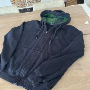Svart hoodie från Polo Ralph Lauren - Svart hoodie från Polo Ralph Lauren med grönt broderat logotyp på bröstet. Utsidan är mjuk och insidan av huvan har ett grönt och mörkblått rutigt foder. Tröjan har snörning i huvan, dragkedja framtill och ribbade muddar vid ärmar och nederkant.