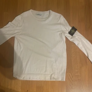 Vit långärmad tröja från Stone Island - Vit långärmad tröja från Stone Island med klassisk rund hals och ikonisk logotyp-patch på vänster ärm. Tröjan är tillverkad i mjuk bomull och har en clean, stilren look som passar perfekt till jeans eller cargo. Snyggt och enkelt plagg för dig som gillar streetwear. Tröjan är i storlek M och har inga särskilda defekter, pris kan diskuteras o skriv för frågor angående tröjan