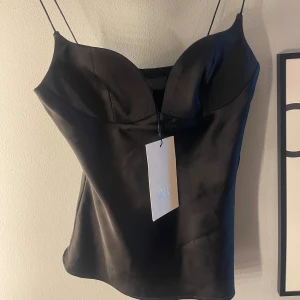 Svart satin korsettopp från Zara - Supersnygg svart korsettopp från Zara i glansigt satinmaterial. Helt ny! - säljer då den var för stor. Toppen har tunna axelband och en markerad byst med djup v-ringning som ger en riktigt trendig look. Perfekt att styla med jeans eller kjol för en cool outfit.
